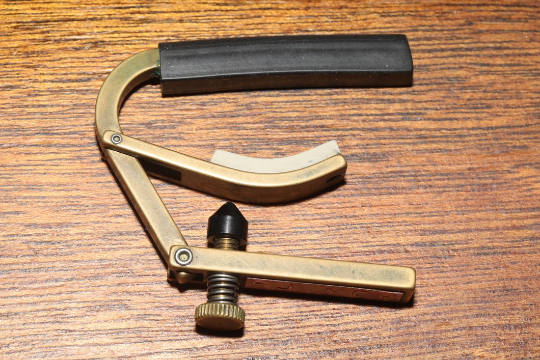 Brass Shubb guitar capo photographed on a wooden surface.
